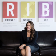 Renee Merrifield smiling and sitting on black couch with T-R-I-B-E posters behind her