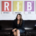Renee Merrifield smiling and sitting on black couch with T-R-I-B-E posters behind her