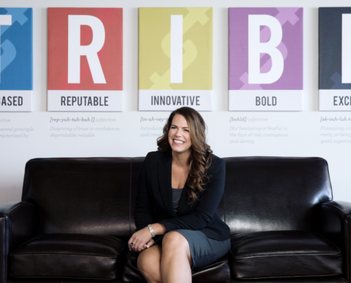 Renee Merrifield smiling and sitting on black couch with T-R-I-B-E posters behind her