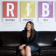 Renee Merrifield smiling and sitting on black couch with T-R-I-B-E posters behind her