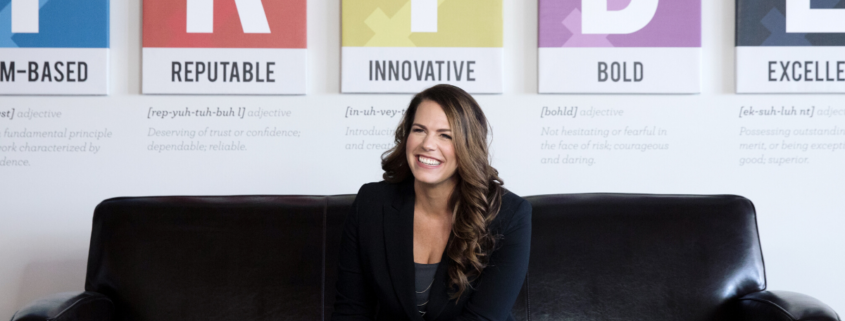 Renee Merrifield smiling and sitting on black couch with T-R-I-B-E posters behind her