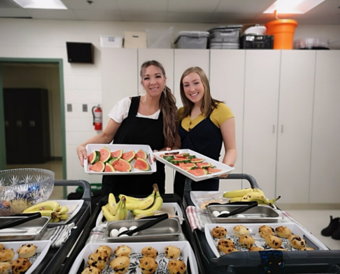 Two women ready to serve breakfast with trays full of eggs, bananas and muffins