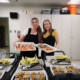 Two women ready to serve breakfast with trays full of eggs, bananas and muffins