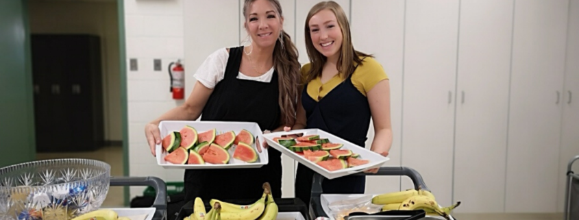 Two women ready to serve breakfast with trays full of eggs, bananas and muffins