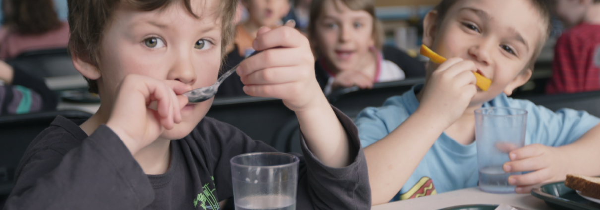 Two kids eating and drinking water with blurred background of other children