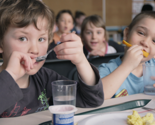 Two kids eating and drinking water with blurred background of other children