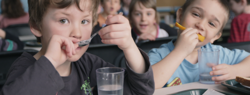 Two kids eating and drinking water with blurred background of other children